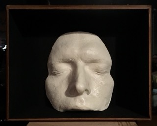 glossy white plaster cast of young man's face capturing forehead, temples, and upper lip, set against a matte black background inside a wooden box